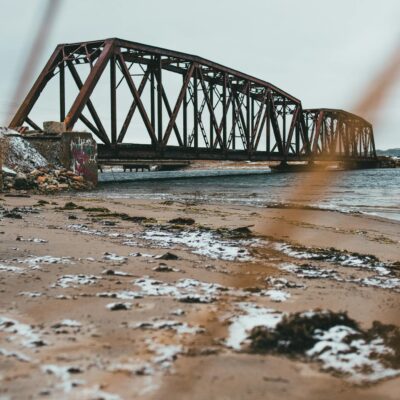 Scenic view of sandy beach with snow against ocean and old bridge under light sky in wintertime