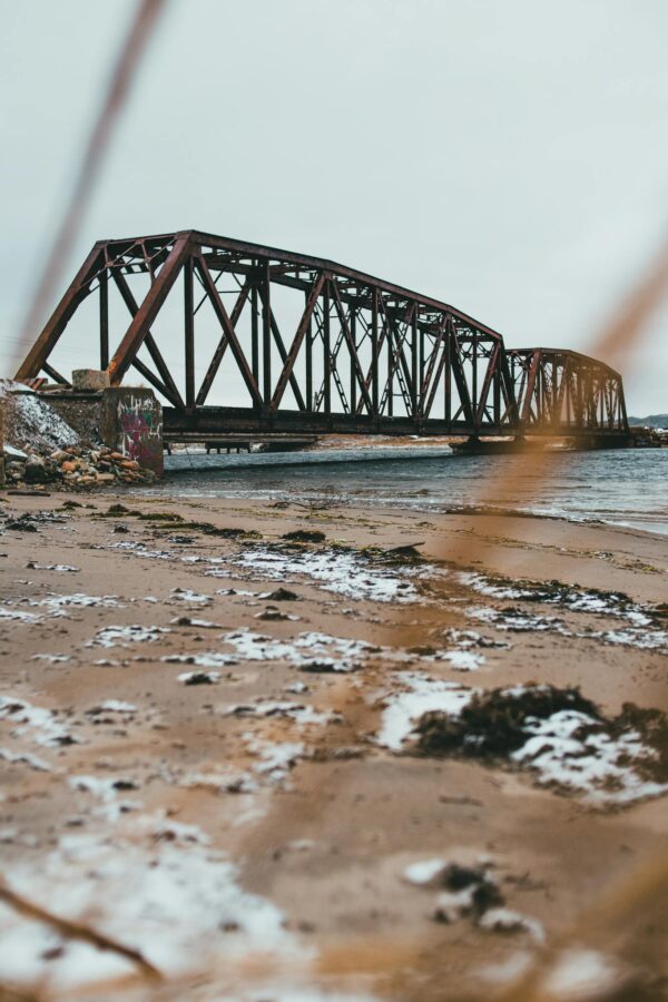 Scenic view of sandy beach with snow against ocean and old bridge under light sky in wintertime